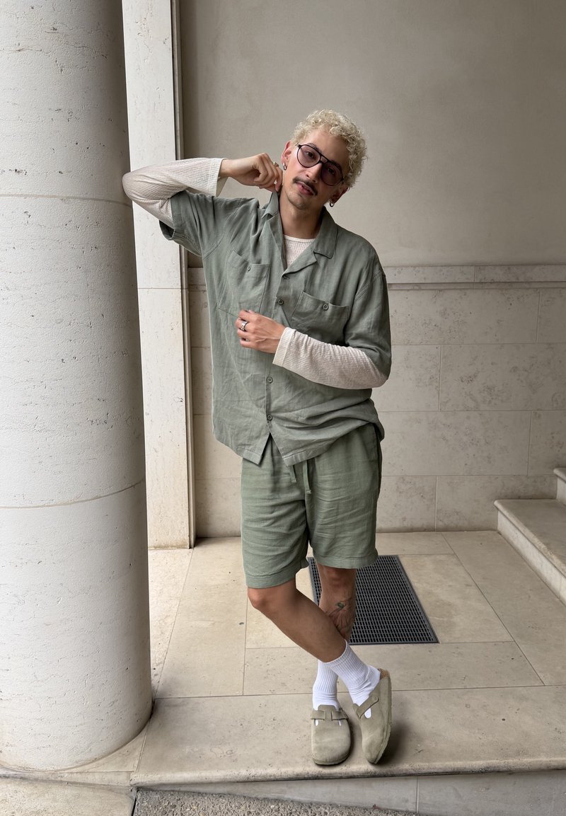Personne aux cheveux clairs bouclés et portant des lunettes, appuyée contre une colonne ronde, vêtue d'une chemise verte à manches courtes, d'un short assorti, d'un haut blanc à manches longues et de chaussons.