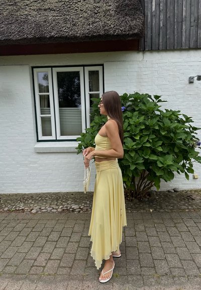 Mujer con un conjunto de dos piezas amarillo y sandalias blancas de pie sobre la acera frente a una casa de ladrillo blanco con un arbusto verde.