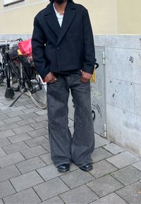 Czarna, oversize'owa marynarka z zaokrąglonymi klapami, w połączeniu z szerokimi, ciemnoszarymi spodniami i czarnymi botkami. Betonowe tło z akcentami rowerów.