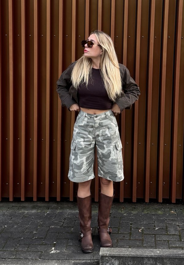 Young woman with long blonde hair wearing a dark crop top, camo cargo shorts, brown jacket, sunglasses, and brown knee-high boots, standing outdoors.