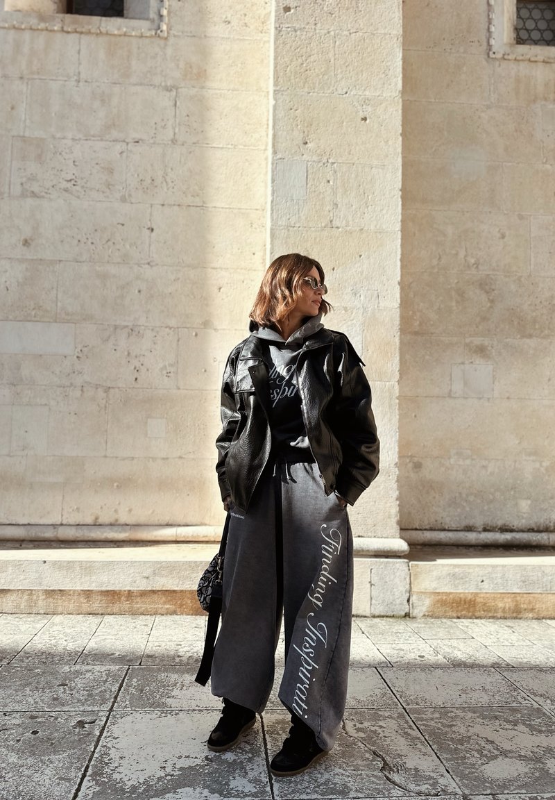 Γυναίκα που στέκεται μπροστά σε πέτρινο τοίχο, φορώντας μαύρο δερμάτινο μπουφάν, γκρι παντελόνι με φαρδιά μπατζάκια και το κείμενο "Finding Inspiriti", γυαλιά ηλίου και μαύρα παπούτσια.