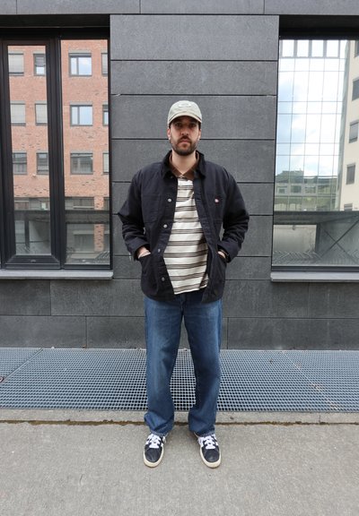 Hombre con gorra beige, chaqueta negra, polo de rayas, vaqueros azules sueltos y zapatillas negras, de pie frente a una pared de piedra gris con ventanas.
