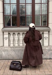 Persoon met lang donker haar, gekleed in een bruine jas en een witte pet, staat voor een groot raam. Een zwarte handtas ligt vlakbij op de grond.
