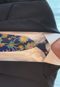Svilena kravata z mornarsko modro podlago in zapletenimi cvetličnimi vzorci v rumeni in zeleni barvi, zavezana okoli ovratnika bele srajce.
