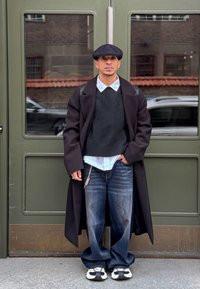 Fekete gyapjúkabát sötét kötött pulóver fölött, világoskék galléros ing, bő szabású farmernadrág és fekete-fehér tornacipő; zöld ajtó mellett állva.