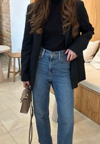 Μαύρο σακάκι πάνω από μαύρο λαιμόκοψη, συνδυασμένο με ψηλόμεσα μπλε τζιν. Κρατώντας μια μικρή μπεζ τσάντα. Άγρια εσωτερική διακόσμηση στο φόντο.