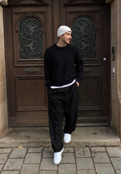 Suéter negro de punto, pantalones negros sueltos y zapatillas blancas. De pie frente a unas puertas de madera ornamentadas con detalles de cristal.