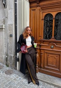 Pantaloni della tuta in raso marrone con strisce dorate, top bianco corto e cappotto nero. Tiene una bevanda verde vicino a una porta in legno con dettagli intricati.