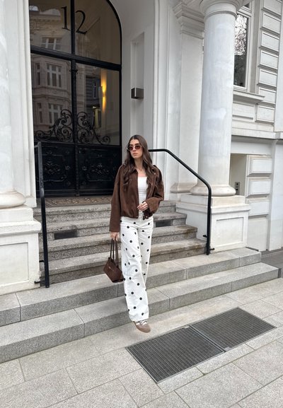 Mujer con chaqueta marrón, pantalones blancos con lunares y gafas de sol bajando unas escaleras de piedra frente a un edificio con grandes puertas negras.