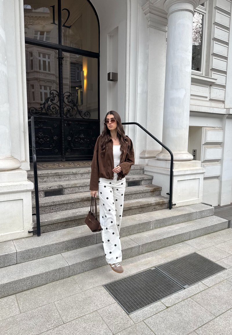 Femeie purtând o jachetă maro, pantaloni albi cu buline și ochelari de soare, coborând trepte de piatră în exteriorul unei clădiri cu uși mari negre.
