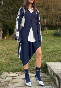 Femme en plein air portant un cardigan Adidas bleu marine et une jupe asymétrique avec une chemise blanche, des chaussettes Adidas, des talons argentés et un petit sac à bandoulière argenté.