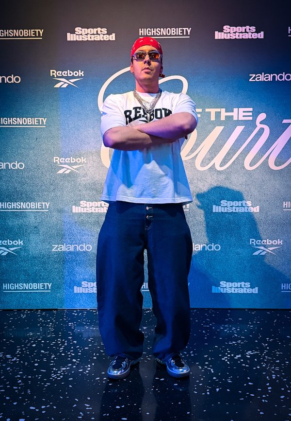 Mann mit rotem Bandana, Sonnenbrille, weißem Reebok-Shirt, blauen Jeans und silbernen Schuhen steht mit verschränkten Armen auf dem Veranstaltungsgelände.