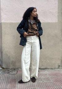 Femme aux cheveux bouclés portant une veste bleu marine, un sweat à capuche marron, un pantalon blanc large et des chaussures marron, debout contre un mur bicolore.