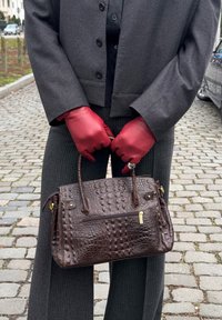 Personne portant des gants en cuir rouges et une veste sombre tient un sac à main marron texturé sur une rue pavée.
