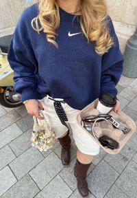 Felpa blu navy con logo Nike bianco, pantaloni corti crema con cravatta a pois nera, stivali marroni fino al ginocchio, che tiene in mano una tazza e un vassoio di oggetti.