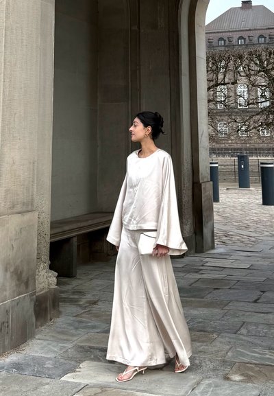 Mujer con un conjunto fluido color crema y sandalias, sosteniendo un pequeño bolso, de pie bajo un arco de piedra con un edificio histórico y árboles sin hojas en el fondo.