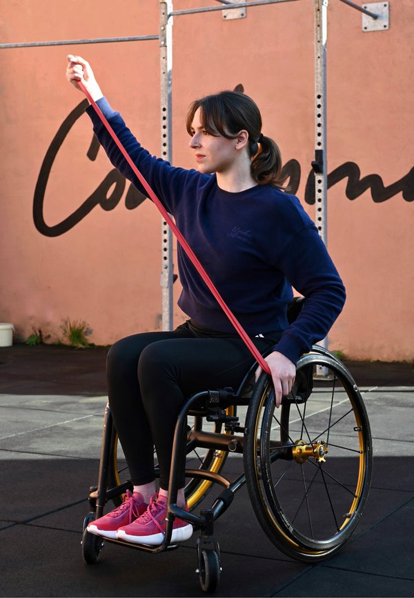 Femme en fauteuil roulant faisant de l'exercice avec une bande de résistance rouge en extérieur, devant un mur couleur pêche avec des inscriptions noires.