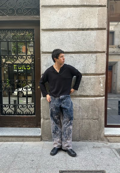 Camisa negra de manga larga, jeans sueltos azul y gris con un patrón de manchas de pintura, combinados con zapatos negros. De pie contra una pared de piedra texturizada.