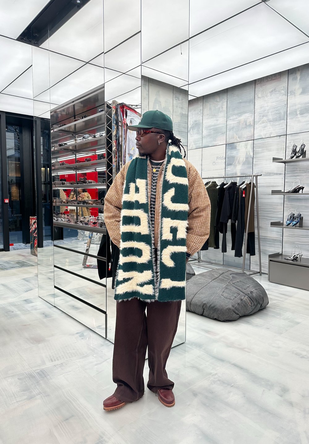 Bufanda verde y crema, chaqueta extragrande de pata de gallo beige, pantalones marrón oscuro y zapatos marrones, exhibidos en un espacio comercial con espejos.