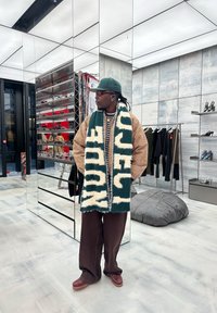 Bufanda verde y crema, chaqueta extragrande de pata de gallo beige, pantalones marrón oscuro y zapatos marrones, exhibidos en un espacio comercial con espejos.