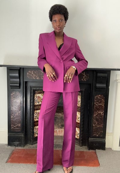 Blazer morado entallado y pantalones de pierna ancha, hechos de una tela suave. El traje presenta solapas afiladas y bolsillos sutiles, complementados con anillos de oro.