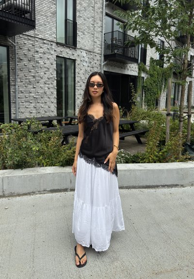 Top sin mangas negro con borde de encaje combinado con una falda blanca fluida y con volantes. La modelo lleva chanclas y gafas de sol elegantes, de pie sobre un suelo de hormigón.