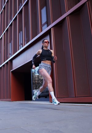 Mujer con ropa deportiva corriendo al aire libre junto a un edificio moderno de metal marrón rojizo, lleva gafas de sol y zapatillas blancas para correr.
