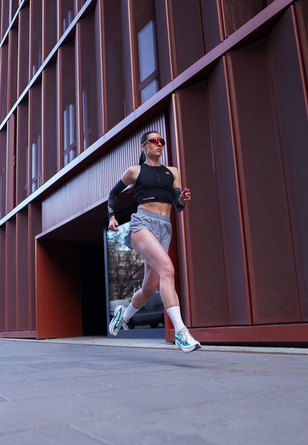 Žena ve sportovním oblečení běží venku vedle moderní červenohnědé kovové budovy, má na sobě sluneční brýle a bílé běžecké boty.