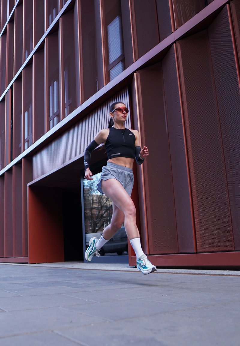 Femme en tenue de sport courant en extérieur à côté d'un bâtiment métallique moderne rouge-brun, portant des lunettes de soleil et des chaussures de course blanches.