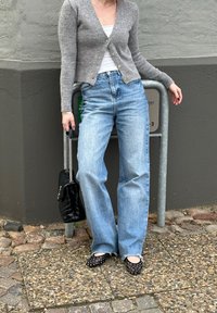 Cardigan grigio chiaro, canotta bianca, jeans a vita alta azzurri chiari, borsa a tracolla nera trapuntata e ballerine nere con pois su una superficie di ciottoli.