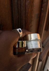 Dva staklenke Estee Lauder drži ruka: tamnosmeđa staklenka sa oznakom "Advanced Night Repair" i svijetlozelena staklenka sa oznakom "DayWear", sjajni metalni poklopci.