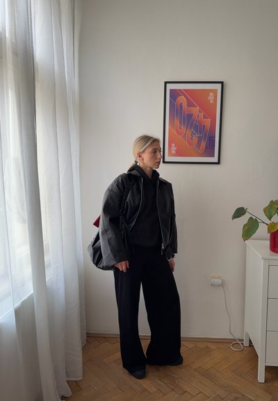 Joven mujer con chaqueta de cuero negra y pantalones anchos está junto a cortinas transparentes en una habitación decorada de forma minimalista con obras de arte enmarcadas y una planta.