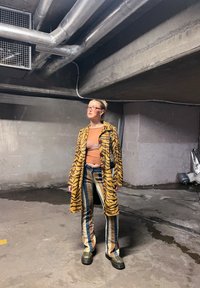 Γυναίκα που στέκεται σε έναν υπόγειο χώρο από σκυρόδεμα, φοράει κίτρινο παλτό με ρίγες τίγρης, ριγέ παντελόνι, πράσινα παπούτσια και γυαλιά, κοιτάζοντας προς τα πλάγια.