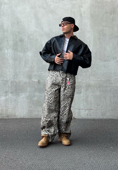 Chaqueta de cuero negra corta, camiseta gris, pantalones oversized de estampado de leopardo y botas de trabajo marrones. Los accesorios incluyen un llavero rosa y gafas de sol.