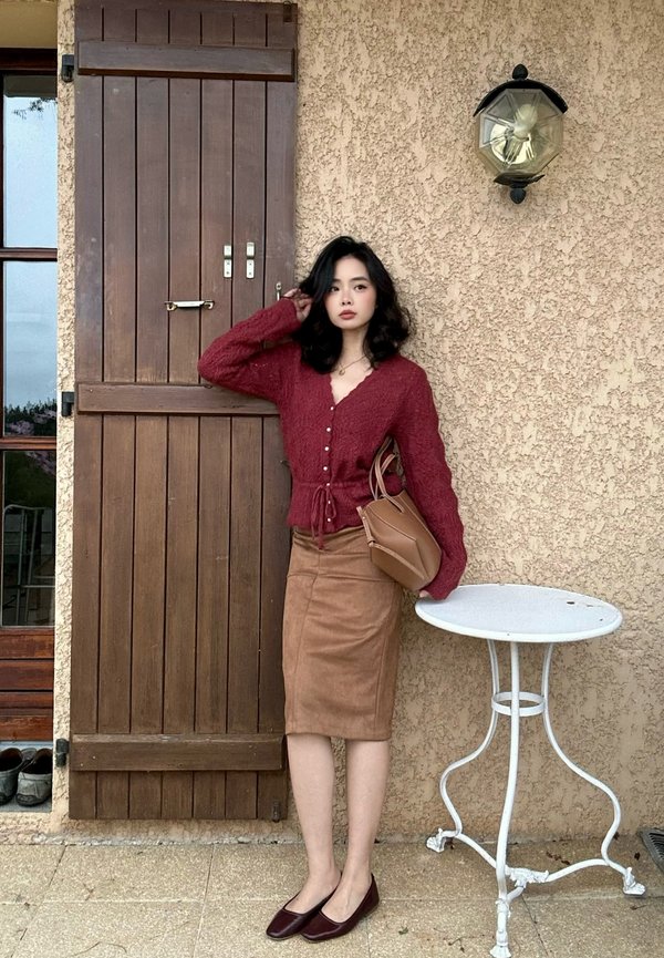 Une femme portant un cardigan rouge et une jupe brune pose à côté d’une petite table blanche, à l’extérieur d’un bâtiment rustique.