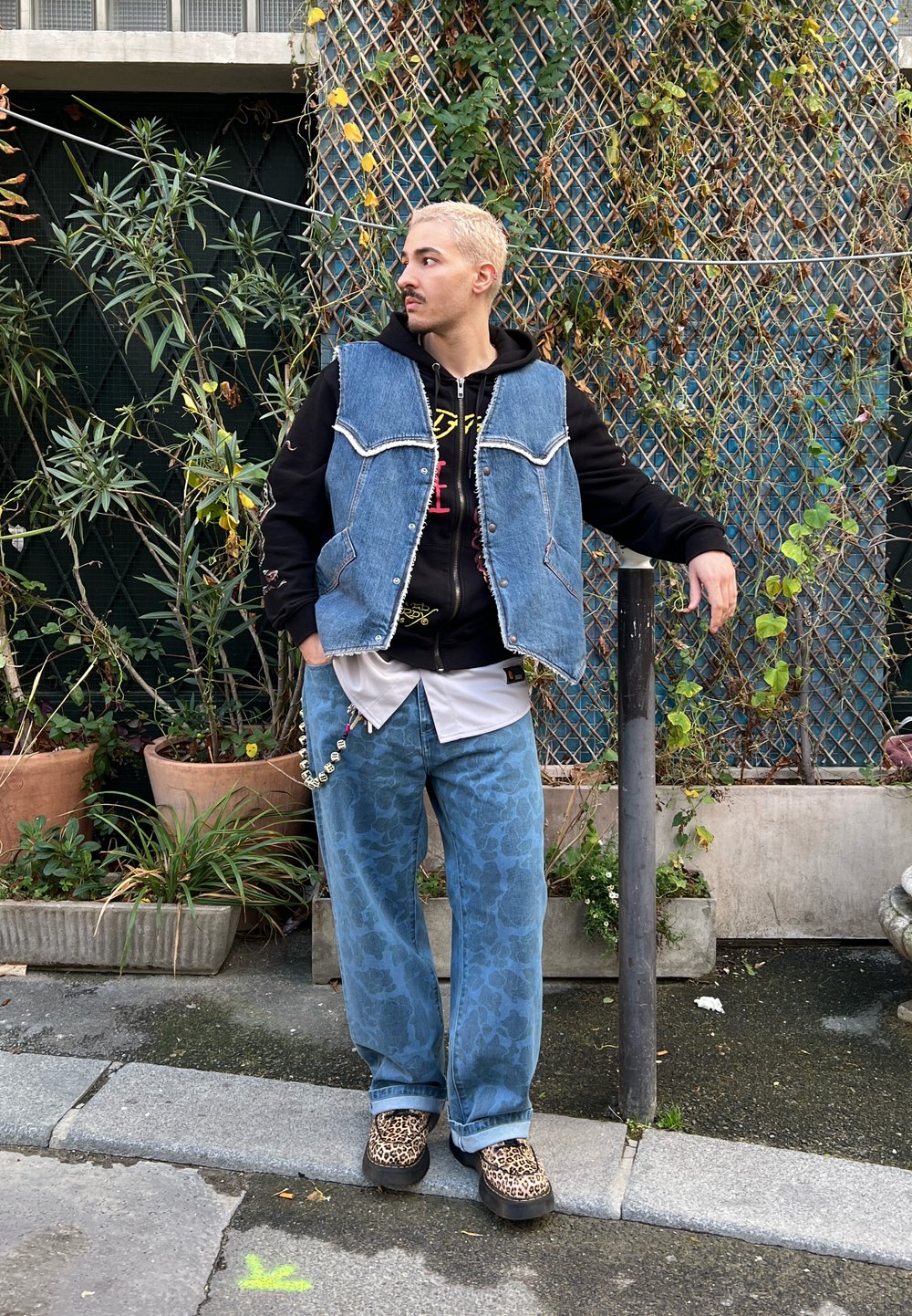 Colete de denim sobre um hoodie preto, combinado com jeans padrão em azul claro e sapatos de padrão leopardo. Os acessórios incluem correntes decorativas.