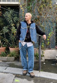 Colete de denim sobre um hoodie preto, combinado com jeans padrão em azul claro e sapatos de padrão leopardo. Os acessórios incluem correntes decorativas.