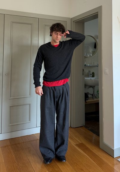 Joven con un suéter negro y pantalones anchos, de pie en el interior sobre un suelo de madera, tocándose la cara cerca del ojo con la mano derecha.