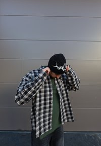 Zwart-wit geplaid overhemd met lange mouwen over een groen T-shirt, gecombineerd met een zwarte beanie met een witte geweefd ontwerp.