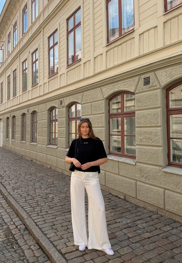 Joven mujer con camisa negra y pantalones blancos está de pie en una calle adoquinada junto a un edificio beige con varias ventanas.