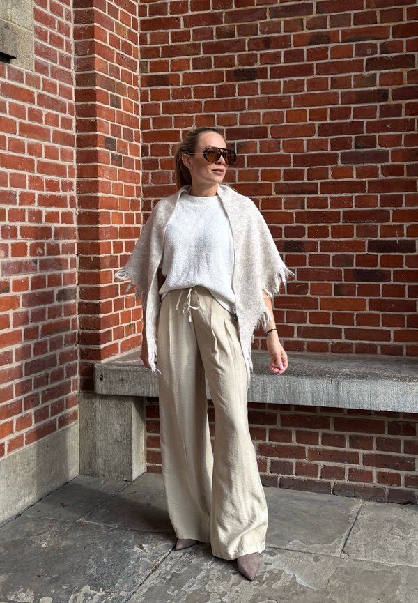 Femeie îmbrăcată în pantaloni bej cu croială largă, top alb și șal deschis la culoare, stând în fața unui zid roșu de cărămidă, cu ochelari de soare și părul strâns în spate.