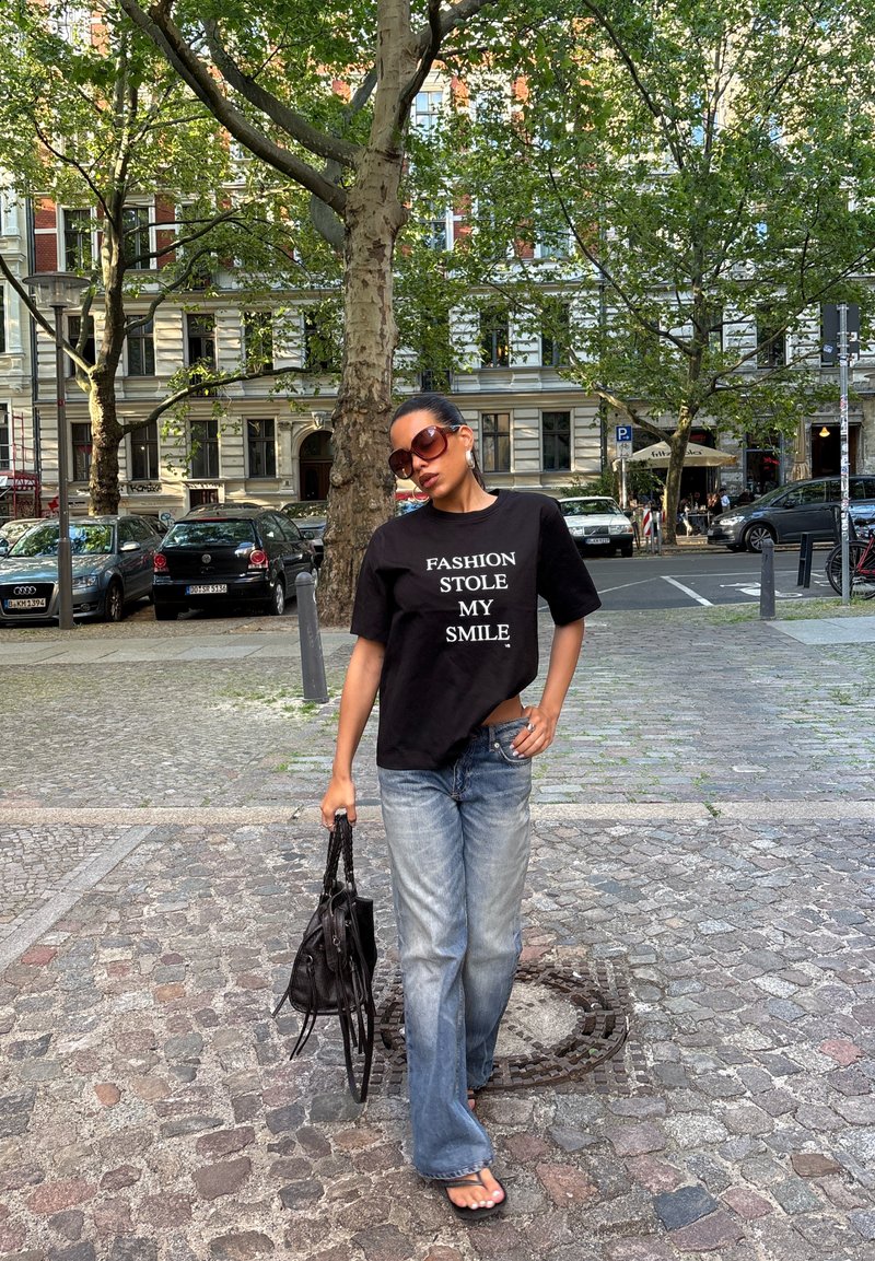 Femme portant des lunettes de soleil et un t-shirt noir avec "FASHION STOLE MY SMILE" marche dans une rue pavée en portant un sac noir.