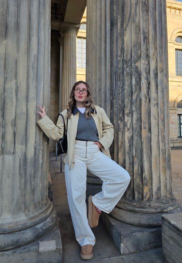 Una persona se apoya en columnas de piedra texturizada, vistiendo una chaqueta crema, suéter gris, pantalones blancos y calzado marrón. Una bolsa negra cuelga a su lado.