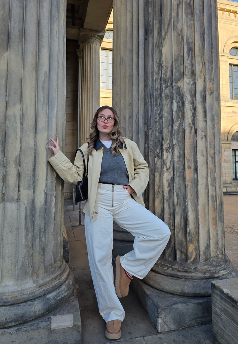 A person leans against textured stone columns, wearing a cream jacket, grey sweater, white trousers, and tan footwear. A black bag hangs at their side.