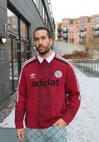 Punane kootud kampsun suure musta "adidas" logoga, valge krae ja sportlikud aktsendid. Omab vertikaalset triibumustrit ja iseloomulikku embleemi.