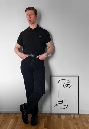 Polo negro de Lacoste, vaqueros negros de tiro alto, zapatillas negras. Dibujo en línea en un marco en el suelo. Suelo de madera y pared gris.