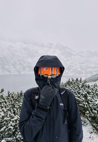 Crna vodootporna jakna s visokim ovratnikom, narančasta prugasta kapa i tamne rukavice. Pozadina prikazuje snijeg i planinsko jezero.