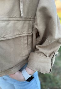 Casaco cropped bege com um tecido suave e texturado, apresentando um meio zíper e punhos abotoados. Usado sobre jeans azul claros e um relógio branco.