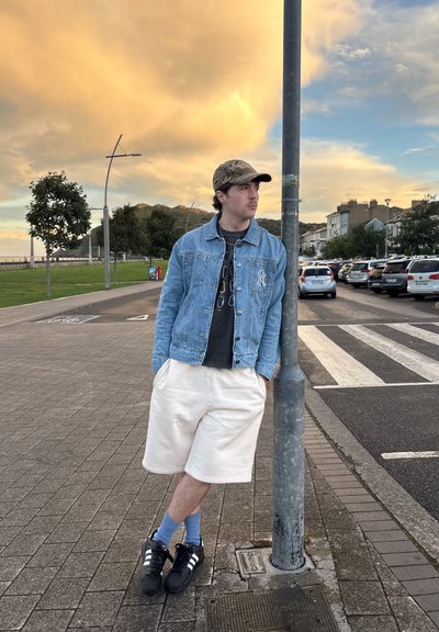 Chaqueta de mezclilla, camiseta gráfica negra, pantalones cortos blancos, zapatos negros con rayas blancas, calcetines azules; apoyado contra un poste en un entorno junto al agua.