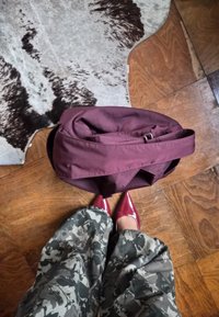 Bordová látková taška s chlopňou a popruhom, položená na drevenej podlahe. V blízkosti sa nachádza vzorovaný šedý a čierny koberec; viditeľné červené topánky.